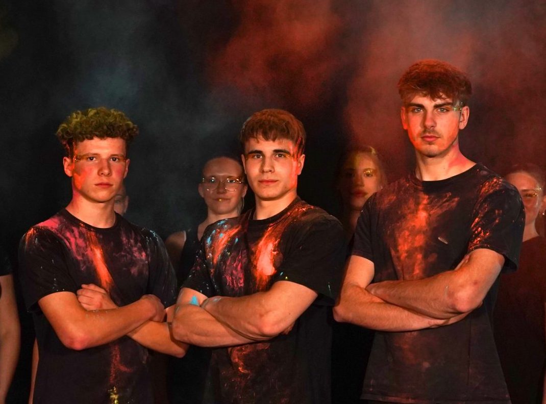 Drei junge Turner mit verschränkten Armen stehen im Vordergrund, in dunkler Kleidung mit orangen Farbpulver-Effekten – weitere Teammitglieder im Hintergrund.