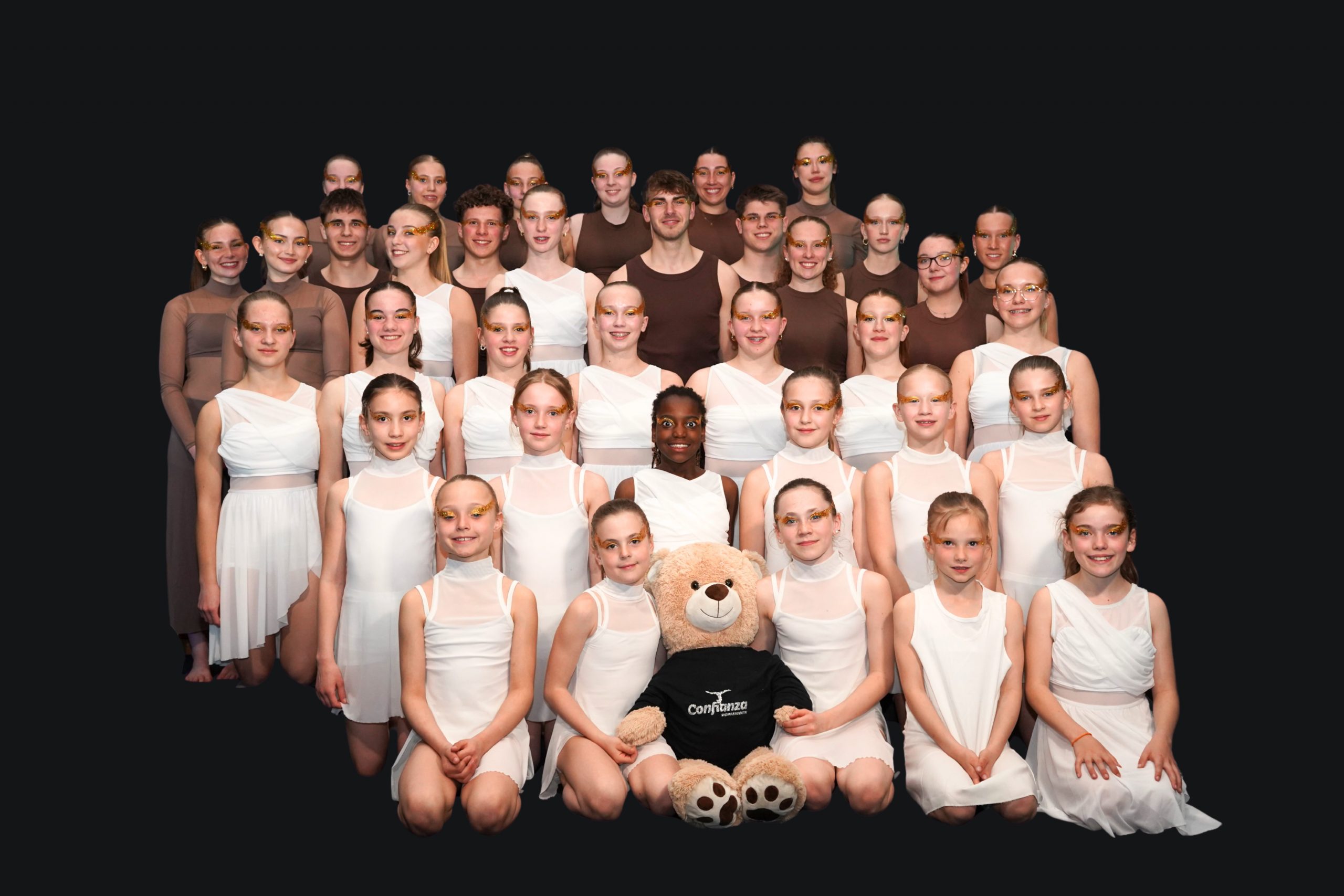 Gruppenbild der Akrobatikgruppe Confianza Westerloy mit Kindern, Jugendlichen und Erwachsenen in weißer und brauner Bühnenkleidung. In der vorderen Reihe sitzt ein Teddybär mit einem Confianza-Shirt.