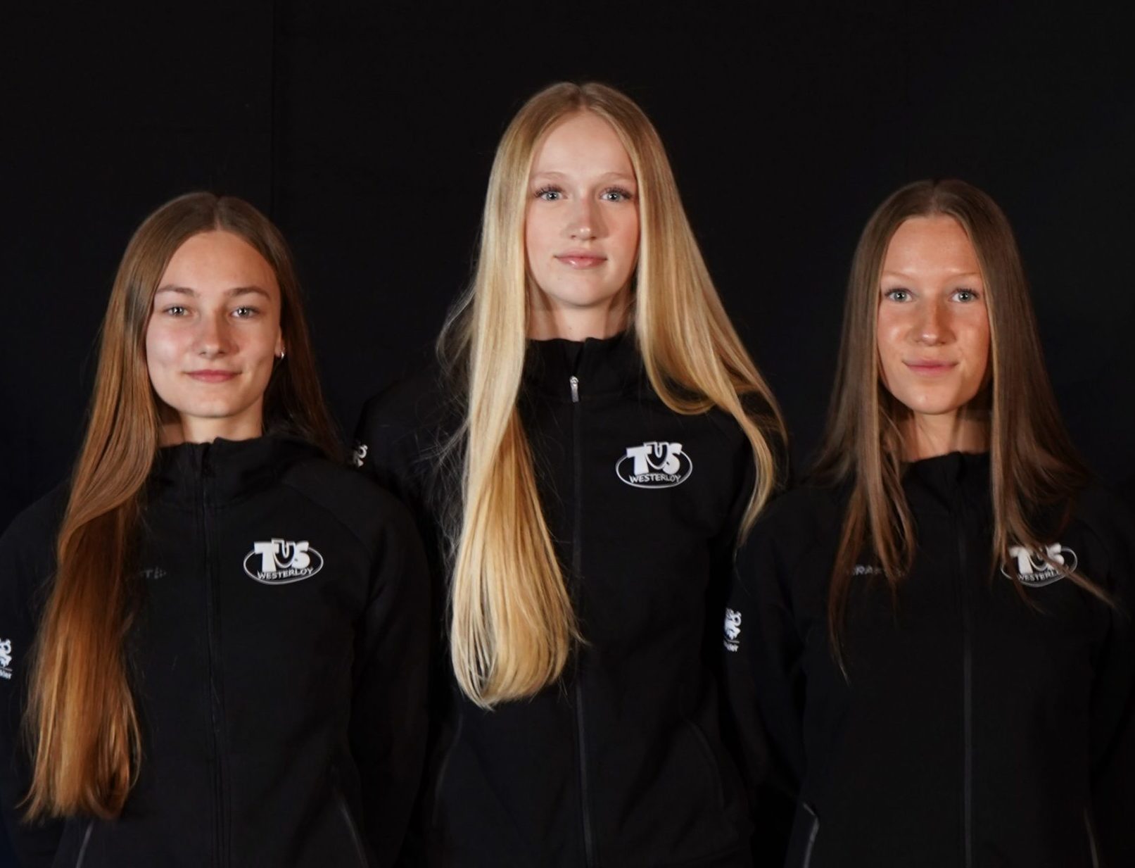 Drei junge Frauen stehen nebeneinander in schwarzen Vereinsjacken mit dem Logo des TuS Westerloy vor schwarzem Hintergrund.