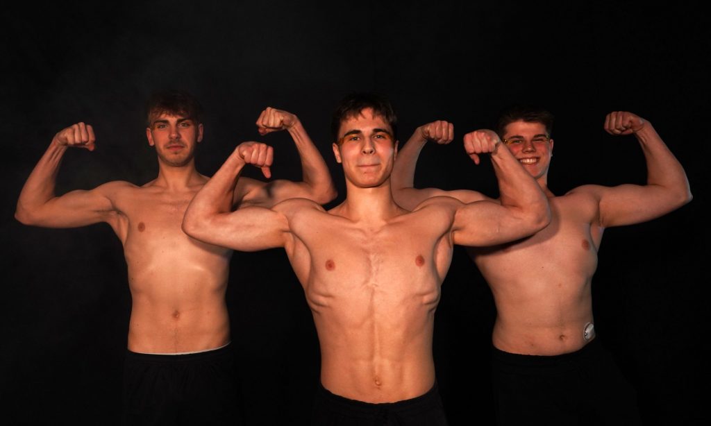 Drei männliche Mitglieder von Confianza posieren oberkörperfrei mit angespannten Armen in Bodybuilder-Pose vor schwarzem Hintergrund