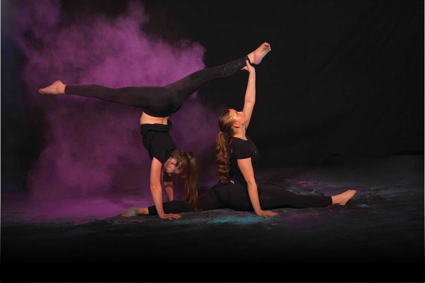 Zwei Turnerinnen in schwarzer Kleidung zeigen eine Bodenakrobatikfigur mit Spagat und Handstand – violetter Farbpulvernebel im Hintergrund.